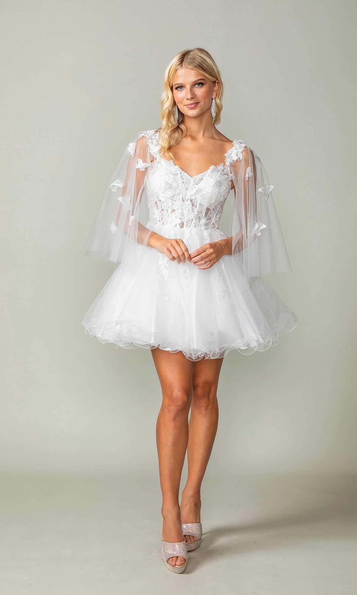 A woman wears the Butterfly-Cape Short A-Line Homecoming Dress 3393, featuring white lace, sheer tulle sleeves, sequin appliques, and beige high heels, standing against a plain light background.